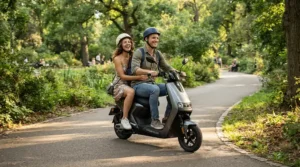 Two adults riding together on a long seat designed for a moped style electric scooter in a park.